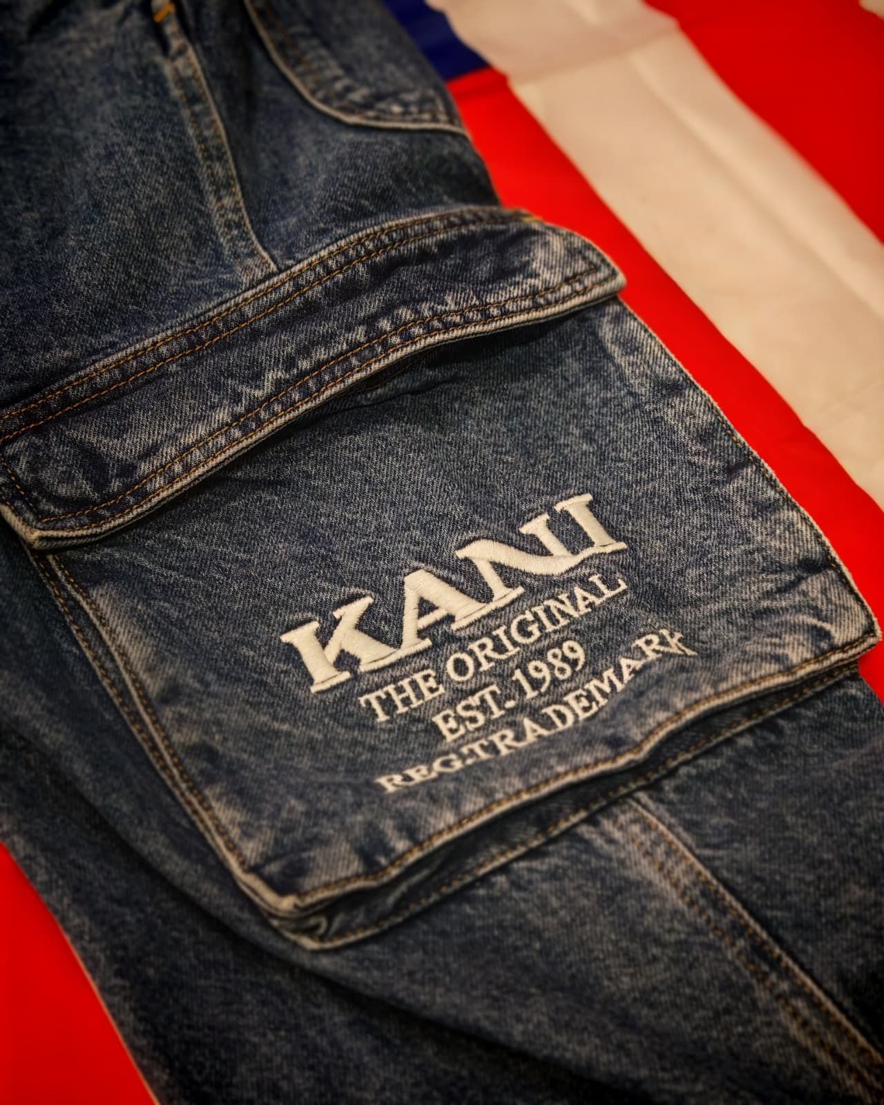 Primo piano di jeans blu scuro con il logo KANU bianco sulla tasca posteriore, accanto a un tessuto rosso e bianco.