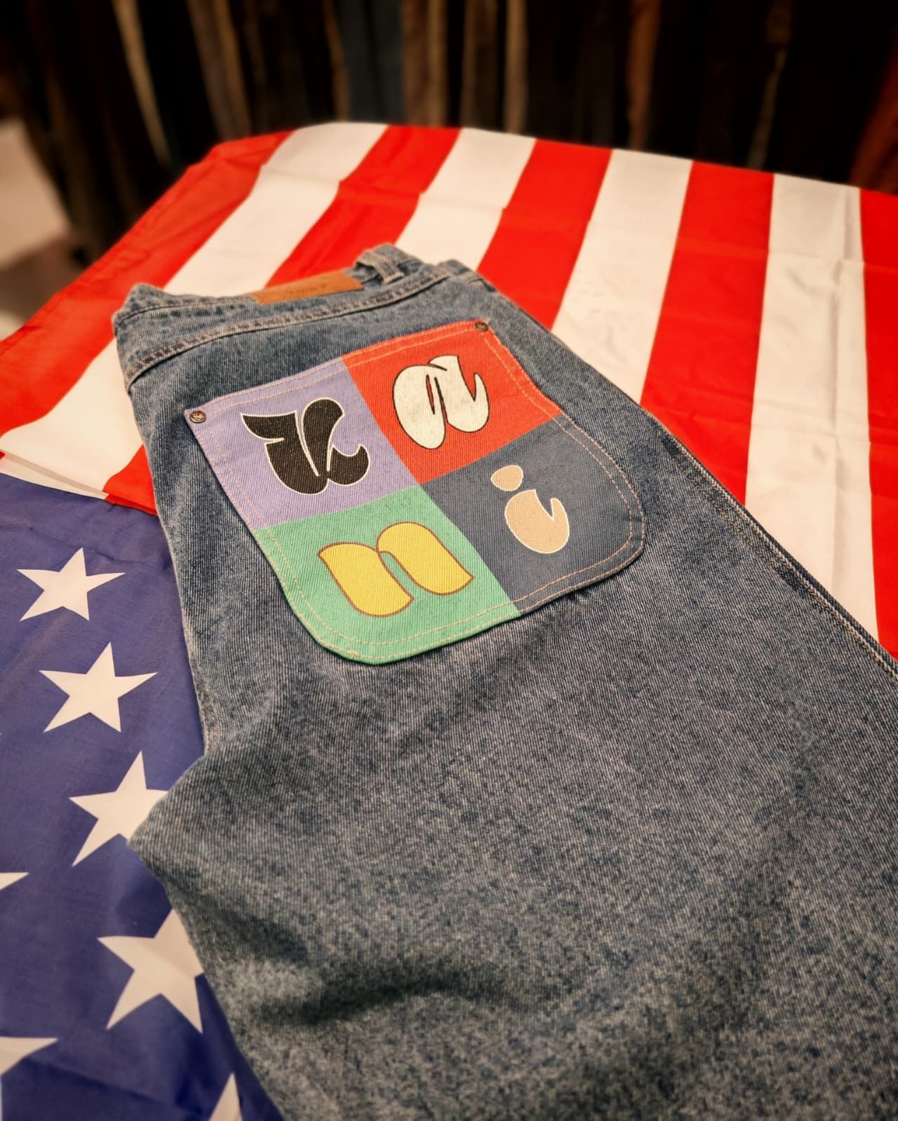 Pantaloni grigi con una toppa colorata a quattro quadranti adagiati su una coperta con la bandiera americana a strisce rosse e bianche.