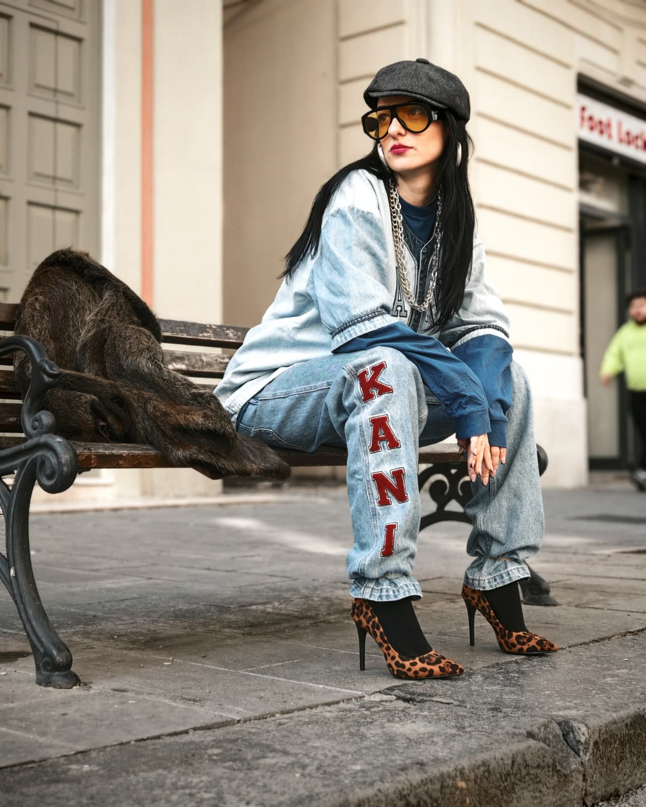 Donna alla moda con occhiali da sole e tacchi leopardati seduta su una panchina in città, con indosso un completo in denim e un berretto.