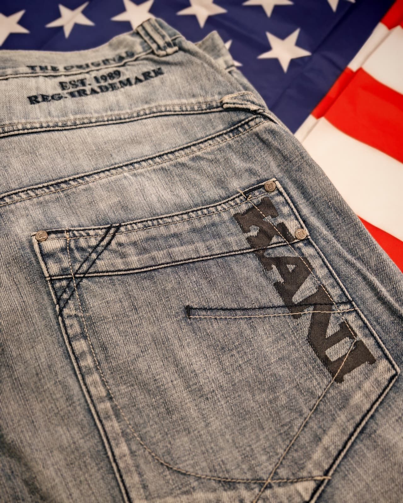 Jeans a lavaggio chiaro con dettagli cuciti sulle tasche, su sfondo con la bandiera degli Stati Uniti.