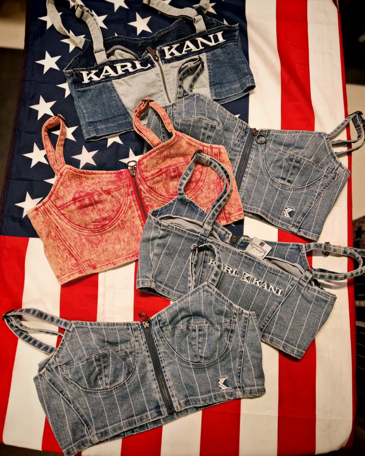Top corti in denim di Karl Kani nei colori blu navy, rosso e grigio su sfondo a righe rosse e bianche.
