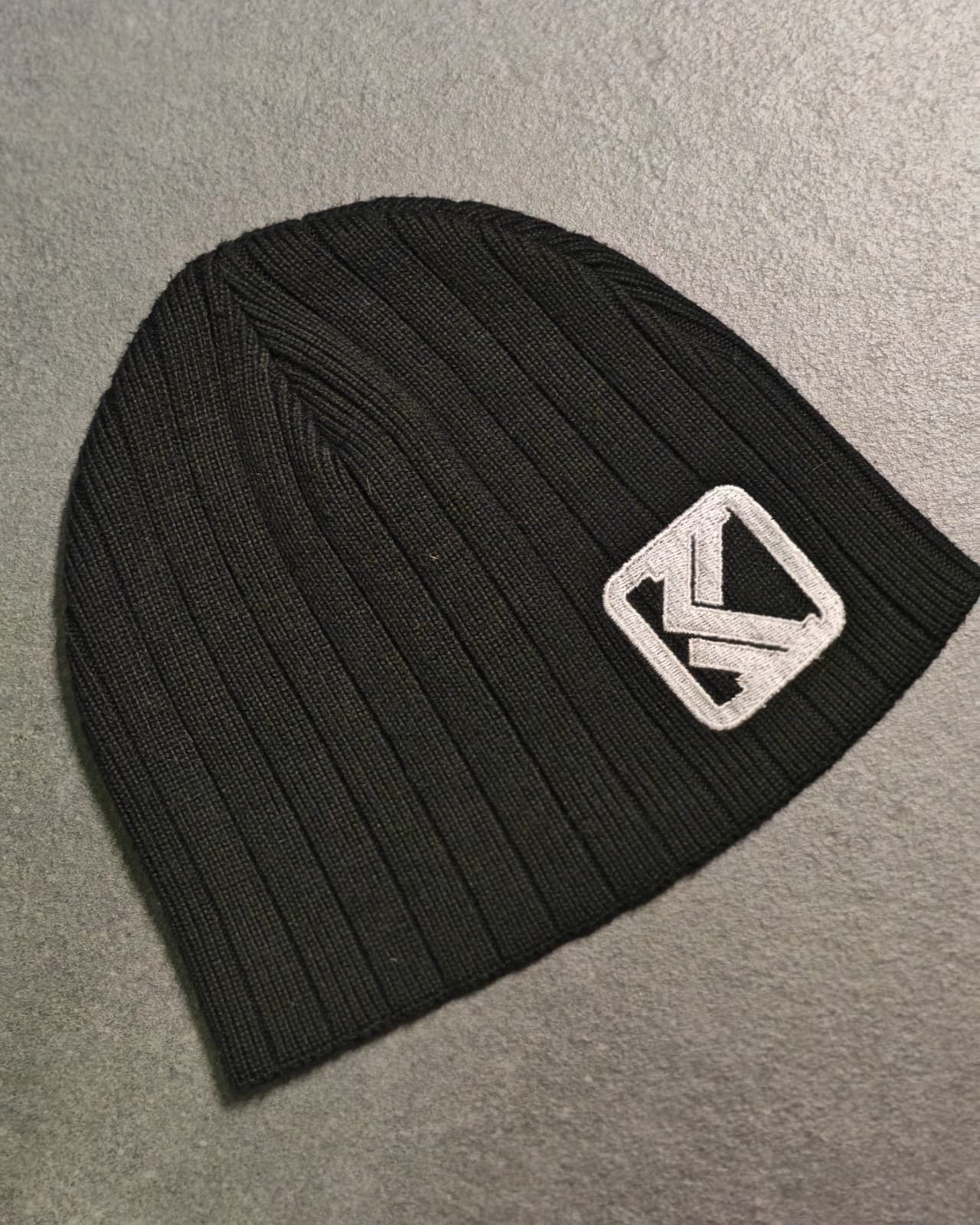 Berretto in maglia nero con logo geometrico bianco su sfondo grigio.