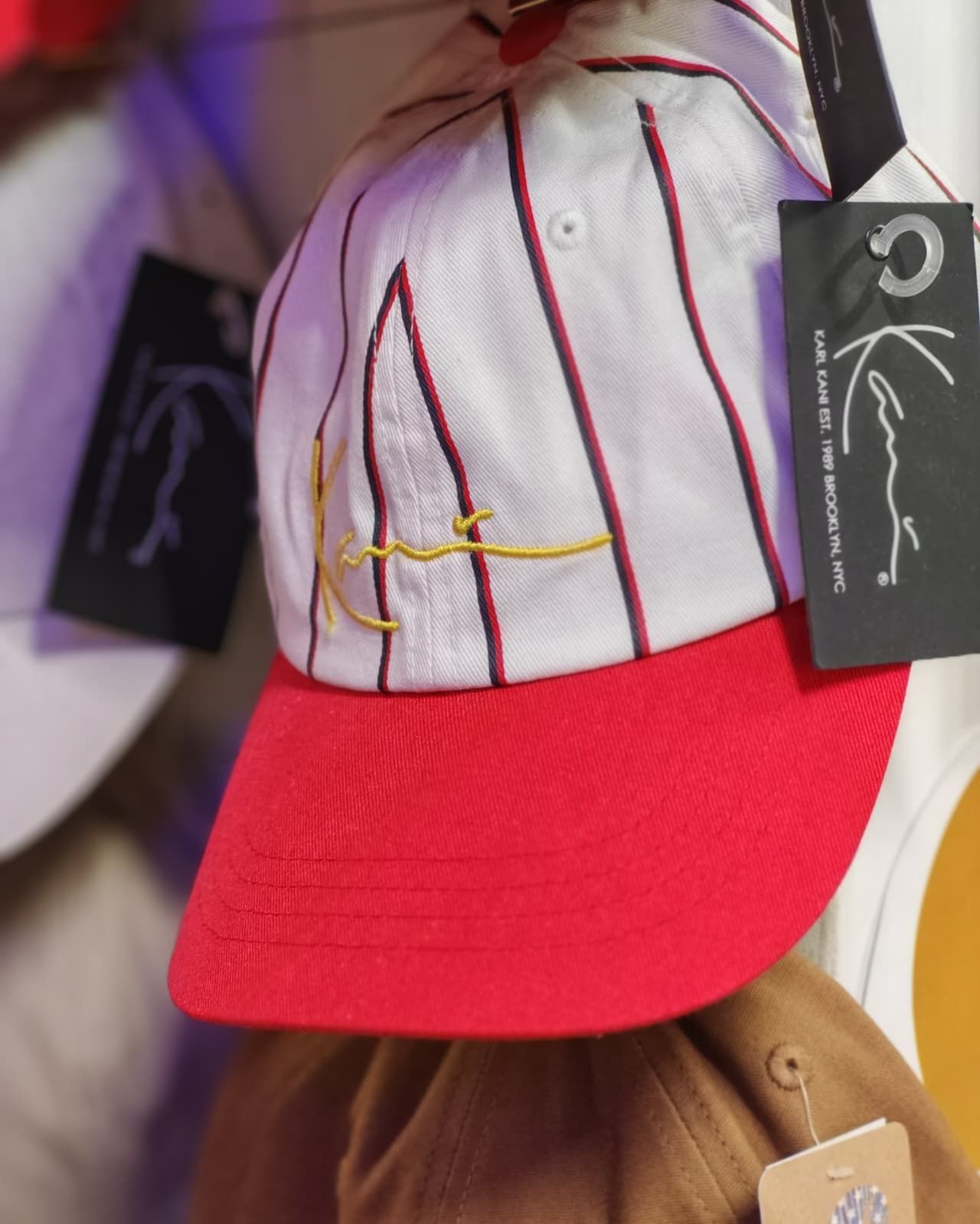 Berretto da baseball bianco e rosso con cuciture dorate e cartellino appeso a un appendiabiti marrone.