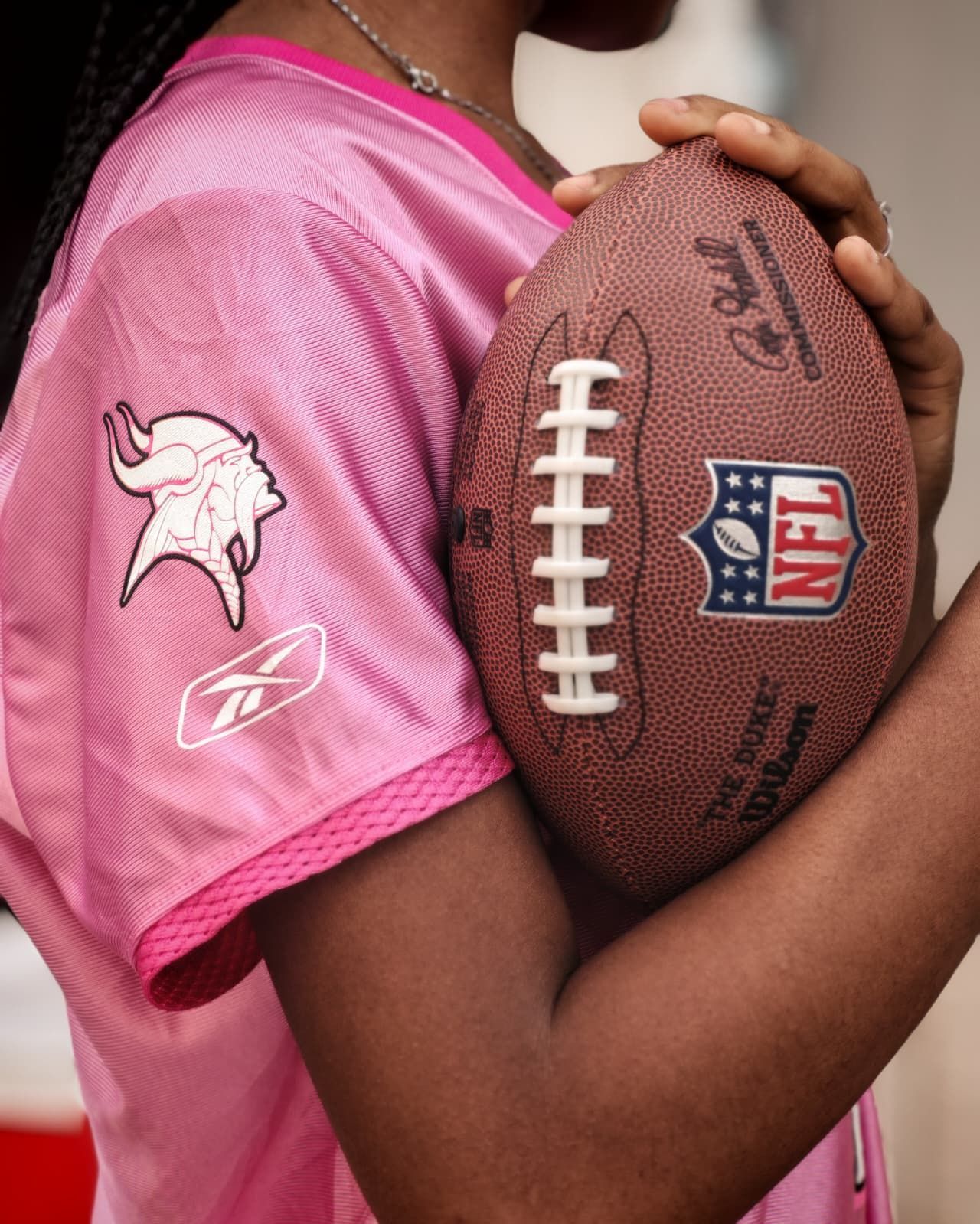 Bambino che tiene in mano un pallone da football americano con indosso una maglia rosa con i loghi della NFL e dei Minnesota Vikings.