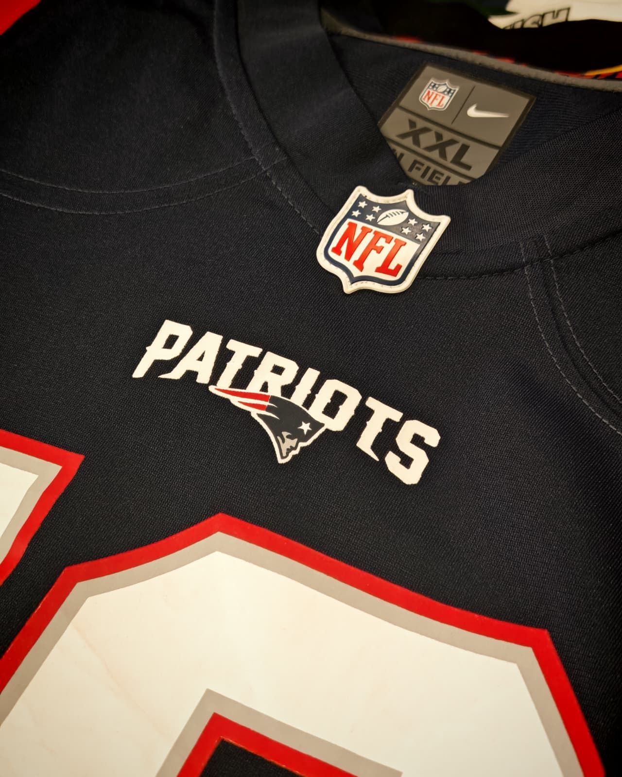 Maglia nera dei New England Patriots della NFL con scritte bianche e rosse, mostrata in primo piano appesa a una gruccia.