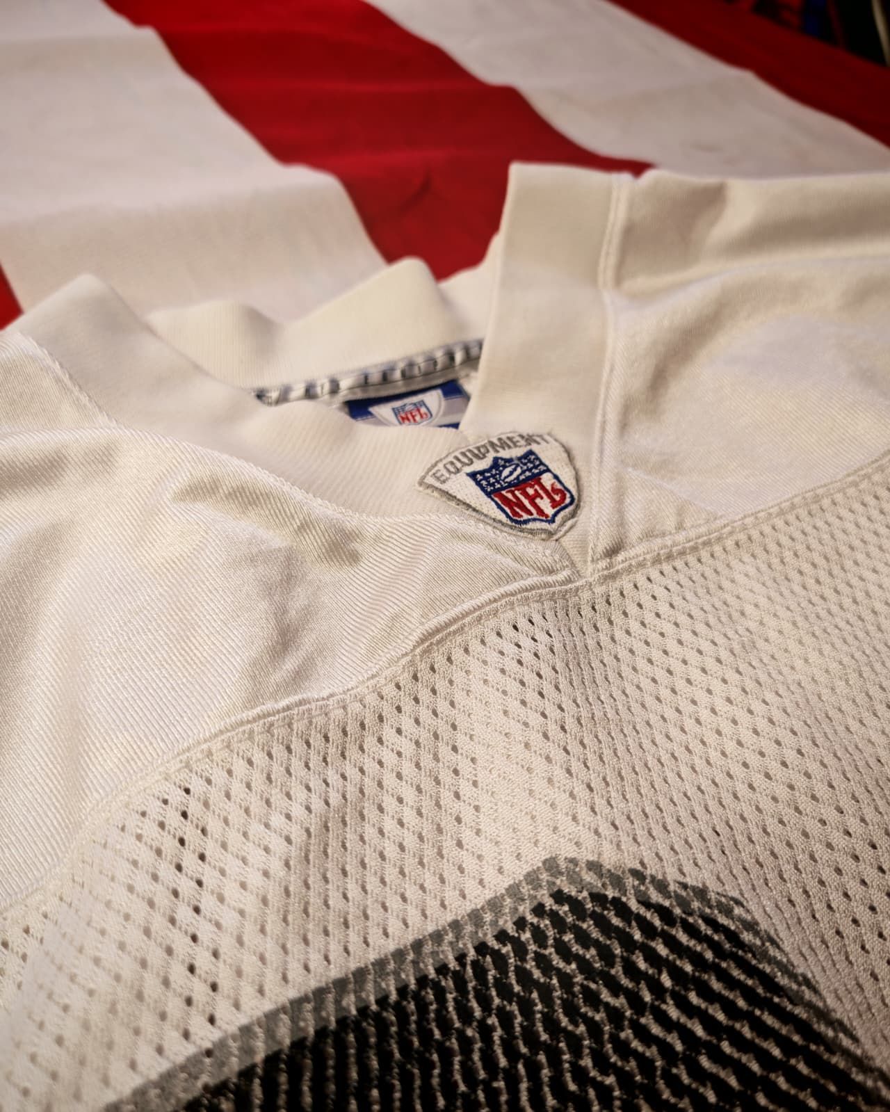 Maglia bianca da football americano (NFL) con uno sfondo a righe rosse e blu e il logo NFL sul colletto.