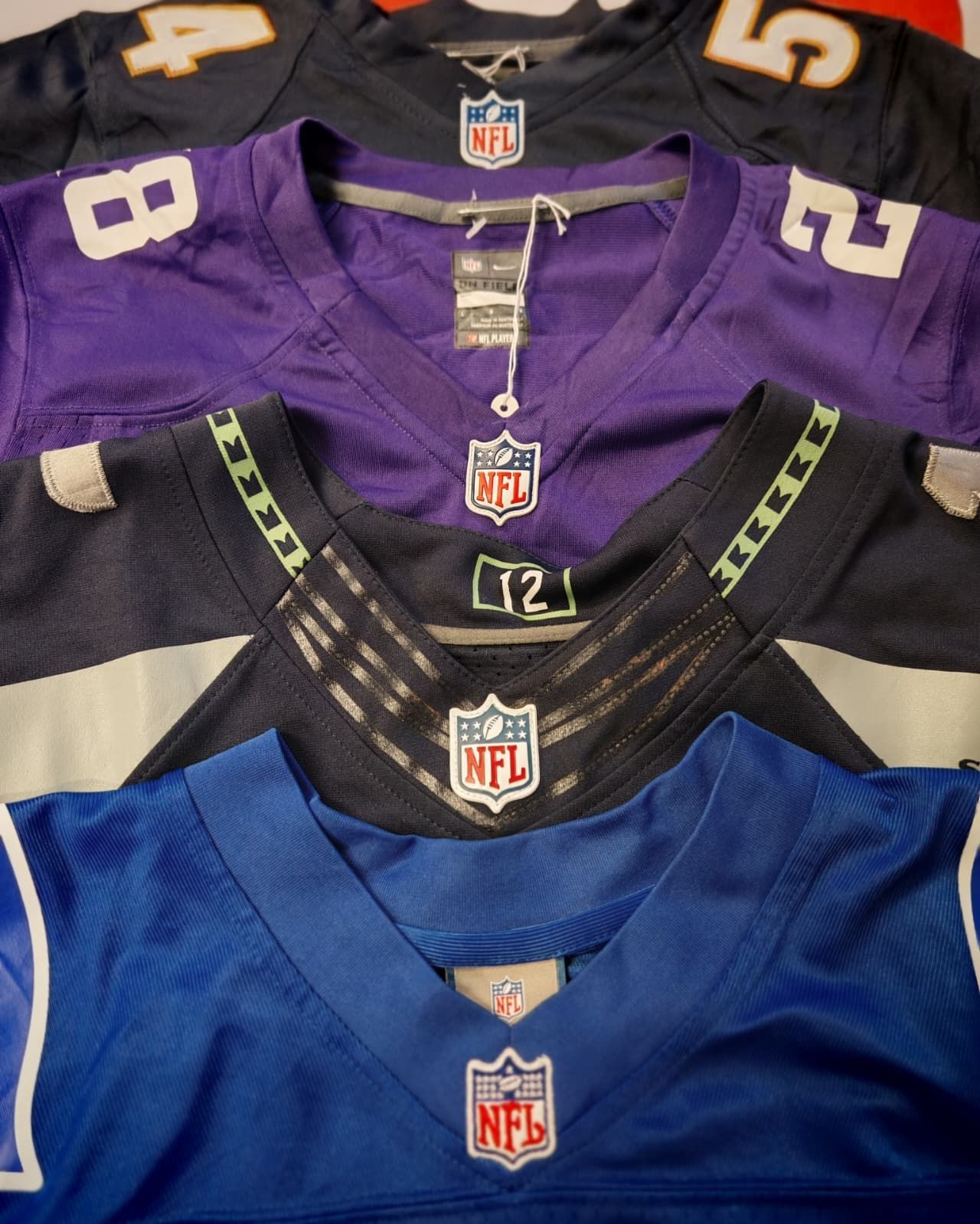 Una pila di maglie da football americano della NFL, nei colori viola, nero, blu e arancione, disposte a strati sovrapposti.