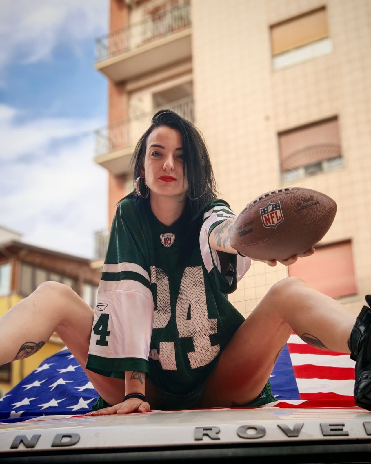 Una donna con indosso una maglia da calcio verde è seduta sul cofano di un veicolo e tiene in mano un pallone da football, con una bandiera americana alle sue spalle.