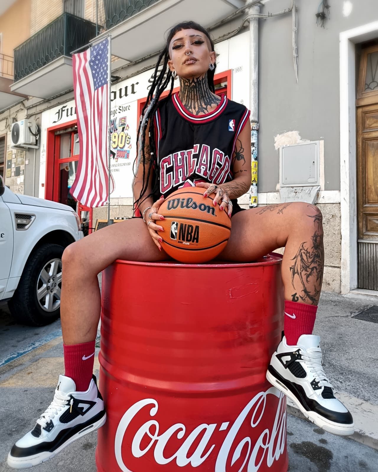 Donna seduta su un barile rosso di Coca-Cola che tiene in mano un pallone da basket, indossa una maglia nera e calzini rossi ed è all'aperto.