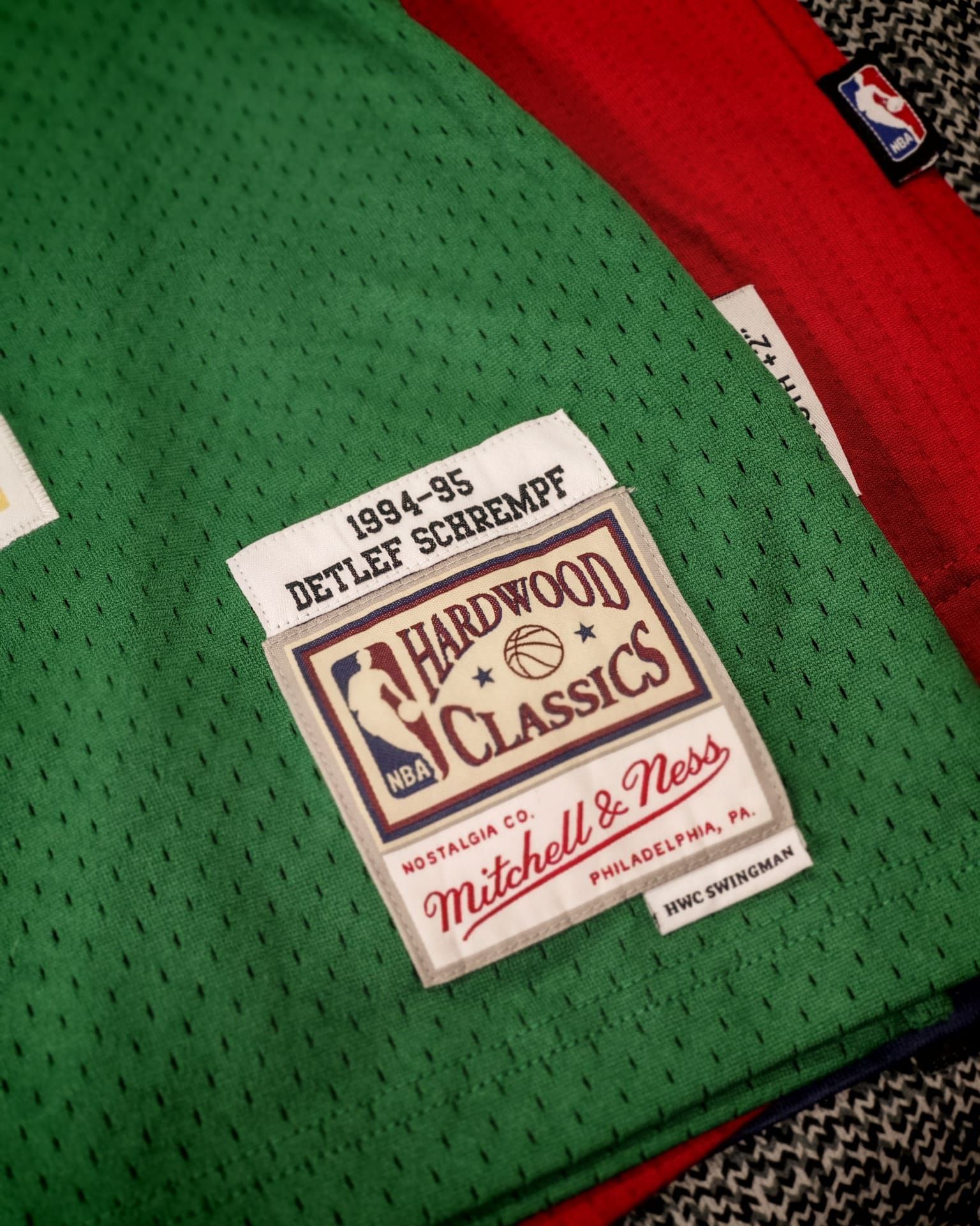 Primo piano dell'etichetta di una maglia Mitchell & Ness Hardwood Classics su tessuto a rete verde con finiture rosse.