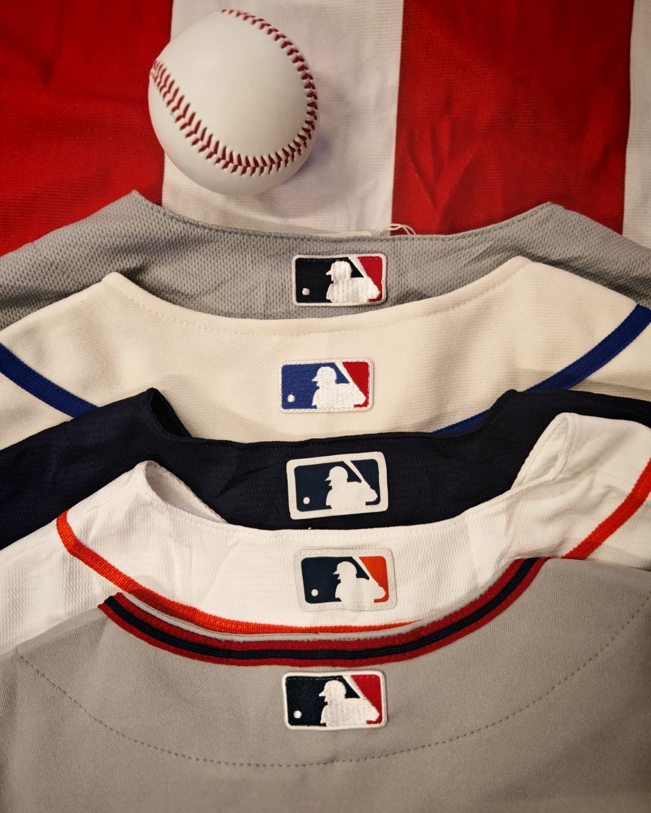 Pila di maglie da baseball della MLB con i loghi, sormontate da una palla da baseball su sfondo rosso.
