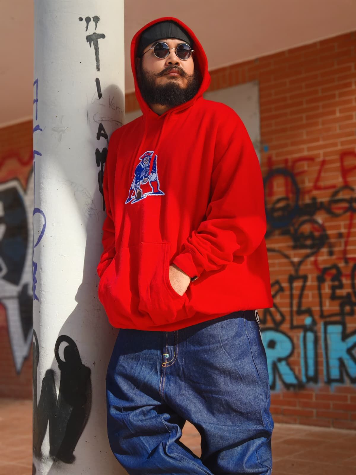 Una persona con una felpa rossa con cappuccio e jeans è appoggiata a un muro ricoperto di graffiti all'aperto.