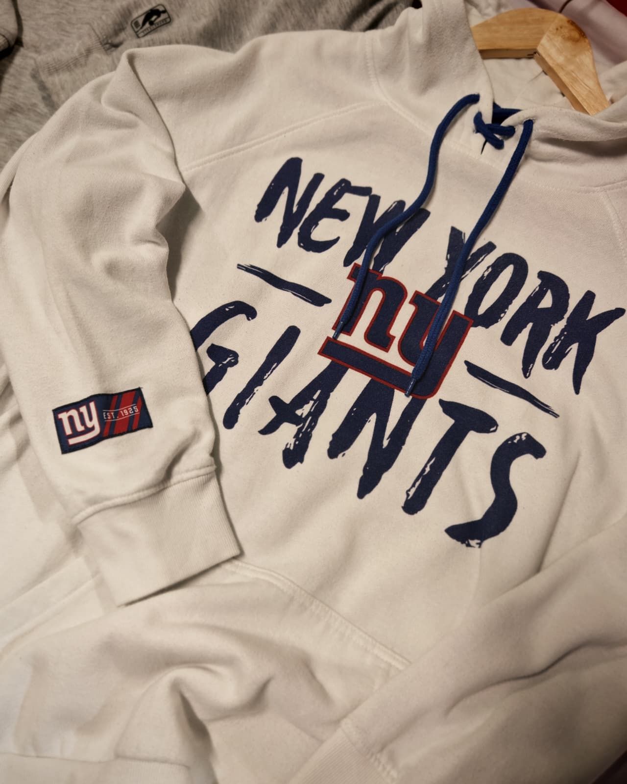 Felpa bianca con cappuccio dei New York Giants con logo blu e rosso, appoggiata su un letto.