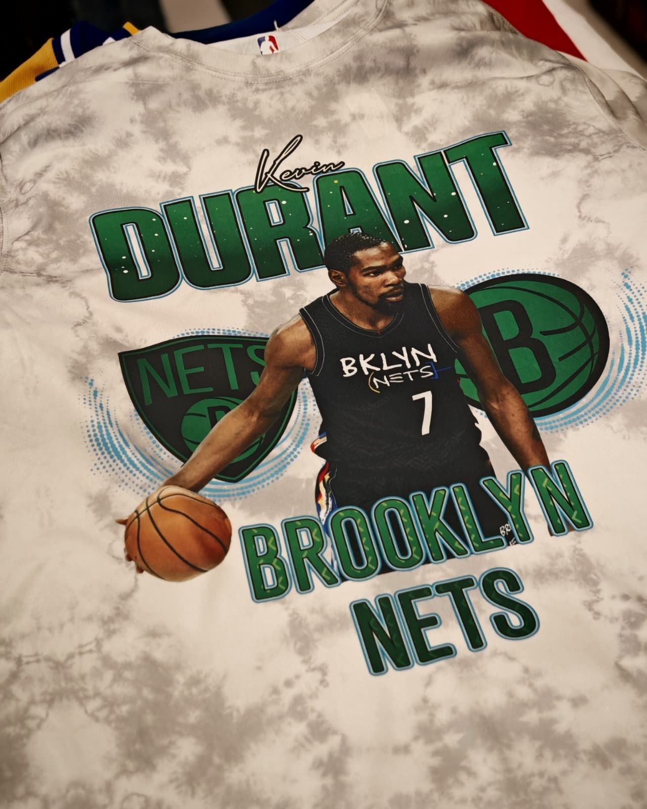 Maglietta bianca tie-dye con grafica di Kevin Durant dei Brooklyn Nets, con pallone da basket e scritte verdi.