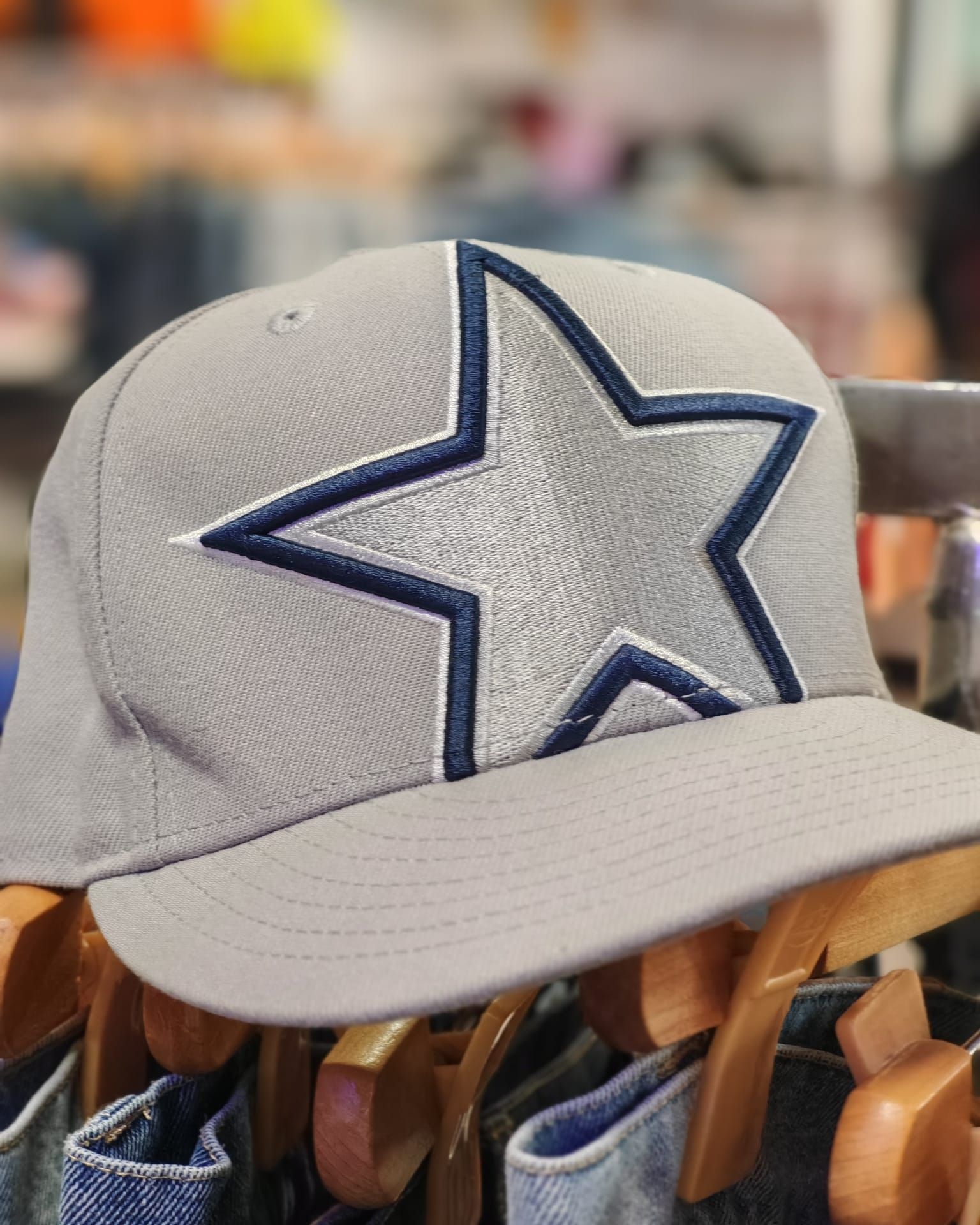 Berretto da baseball grigio con una stella bordata di blu scuro sul davanti, appeso a grucce di legno in un negozio.