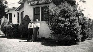 Two men stand outside a mortuary building. The building has a sign and a large window.