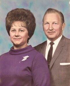 Couple posing for a portrait. Woman in purple dress, brooch; man in gray suit, tie. Blue background.