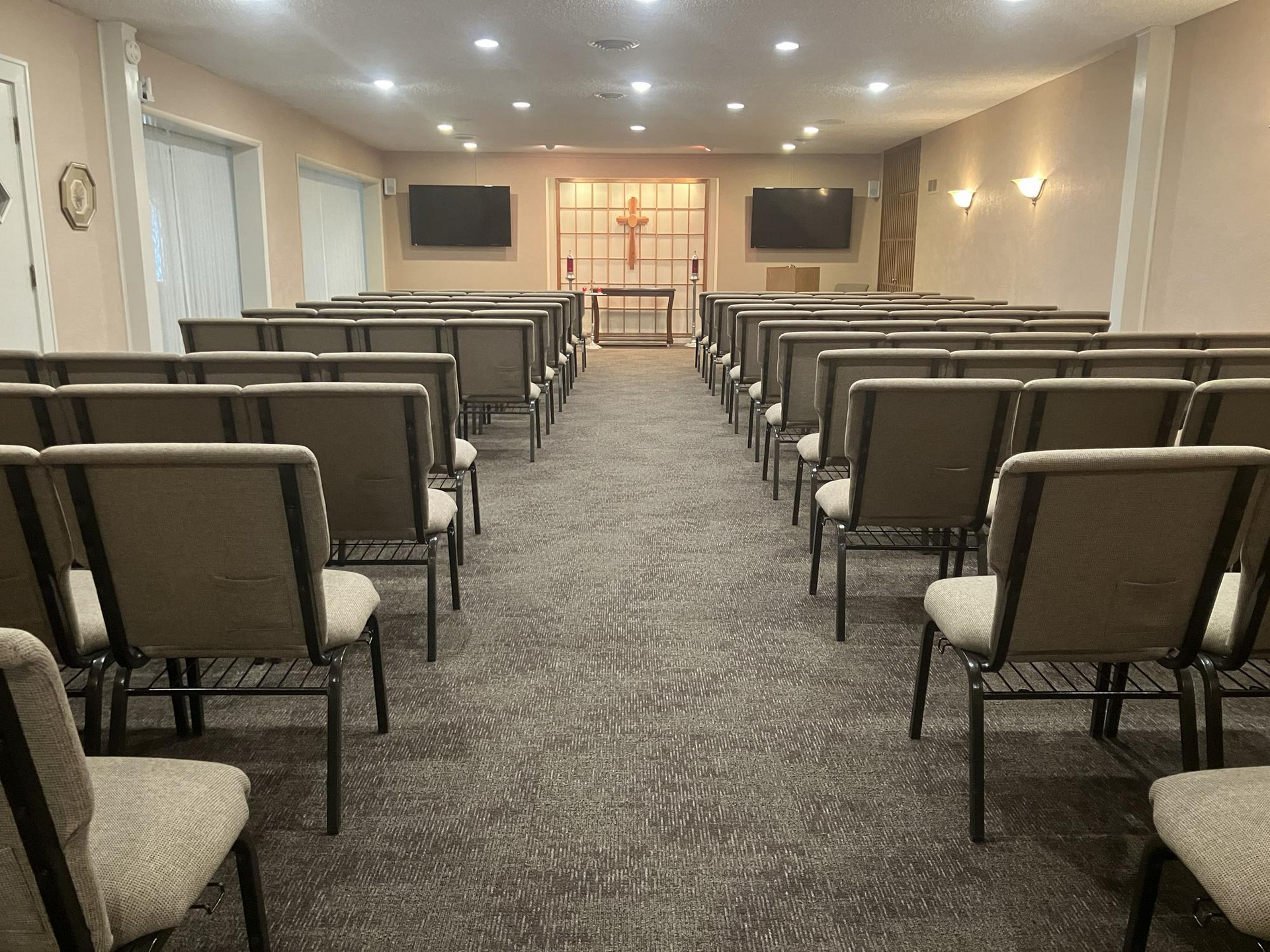 Rows of chairs in a chapel, facing an altar with a cross. Two TVs hang on the wall.