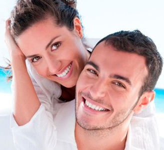 Smiling couple embracing; woman behind man, both in white, outdoors.