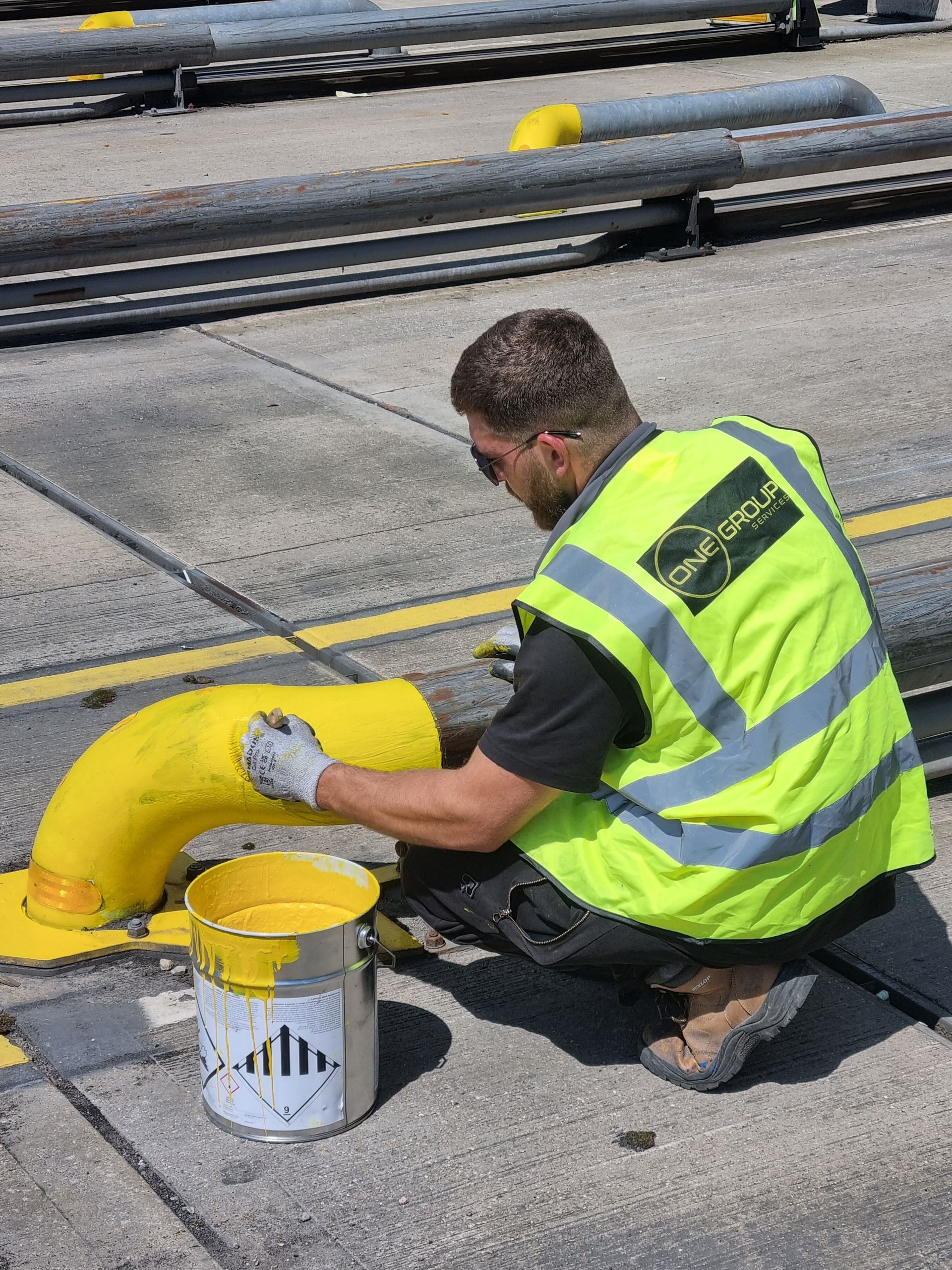 A man in a yellow vest is kneeling down and painting a yellow pipe.