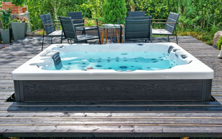 A rectangular hot tub with blue water on a dark wooden deck, surrounded by patio furniture and greenery.