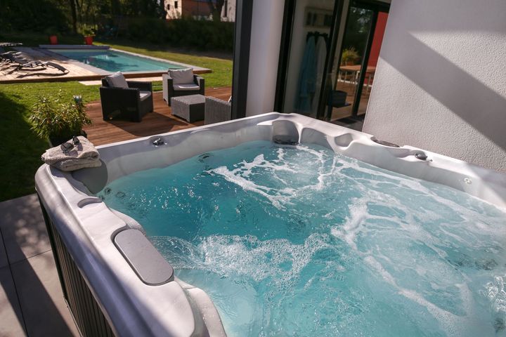 An outdoor hot tub with bubbling blue water on a patio, with a swimming pool and patio furniture in the background.