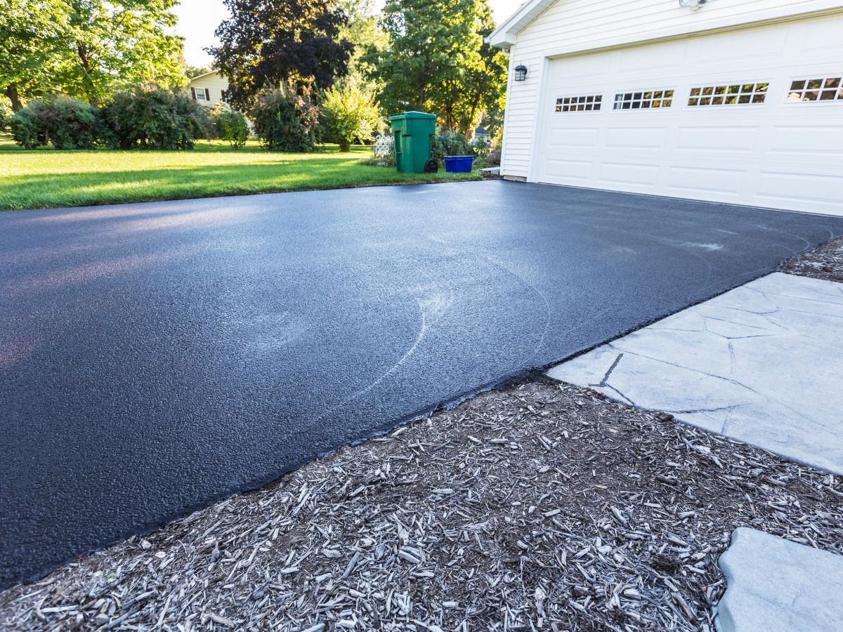 Freshly paved black driveway beside a white garage, with grass and trees in the background
