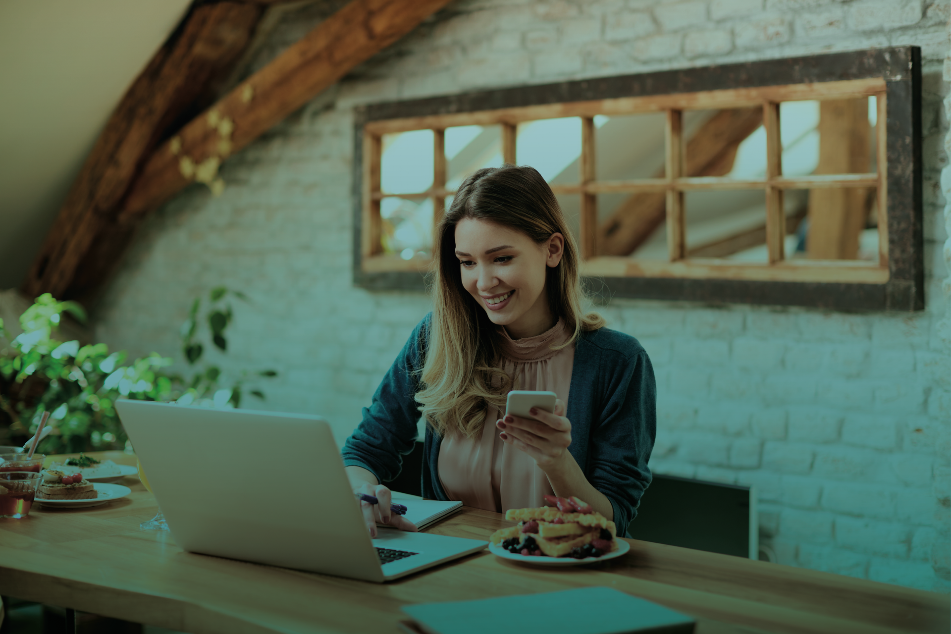Uma mulher está sentada à mesa usando um laptop e um celular.