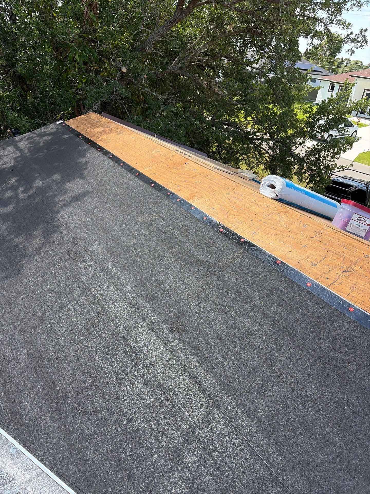 Roof with dark shingles and tan underlayment, with trees and a few tools at the edge.
