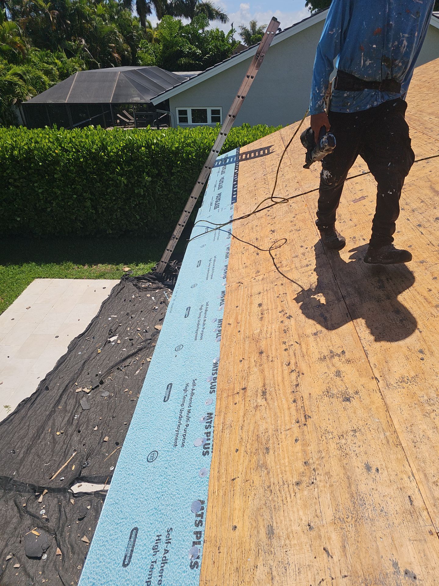 Worker on a roof installing blue underlayment along the edge of a shingled roof.