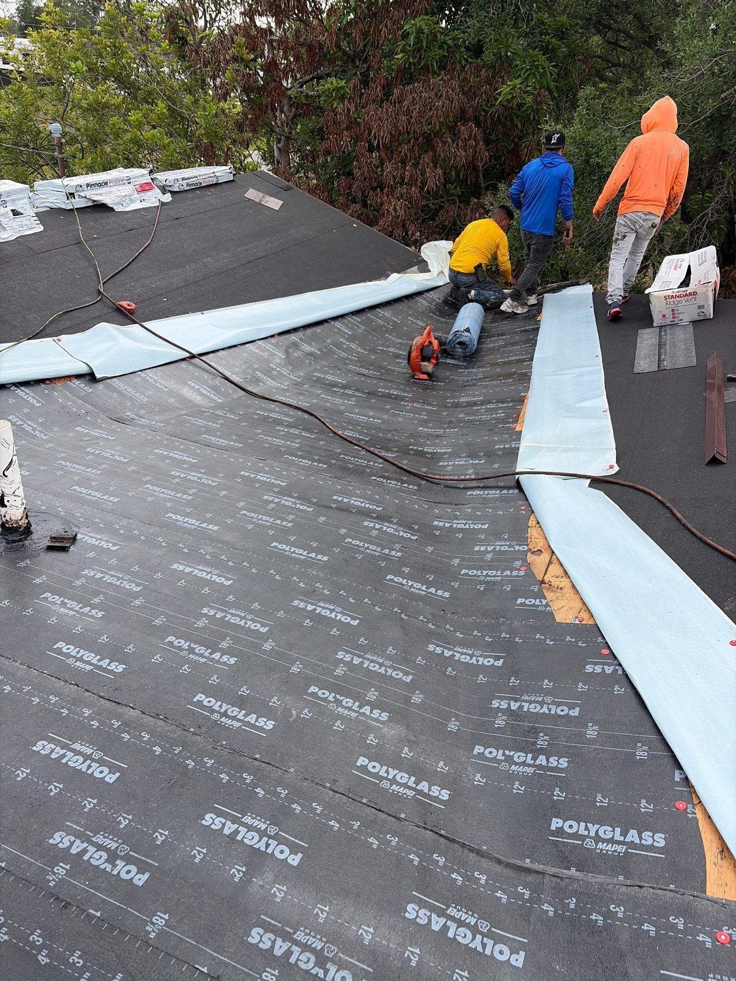 Workers installing roofing underlayment on a sloped roof, with blue and orange rain jackets visible.