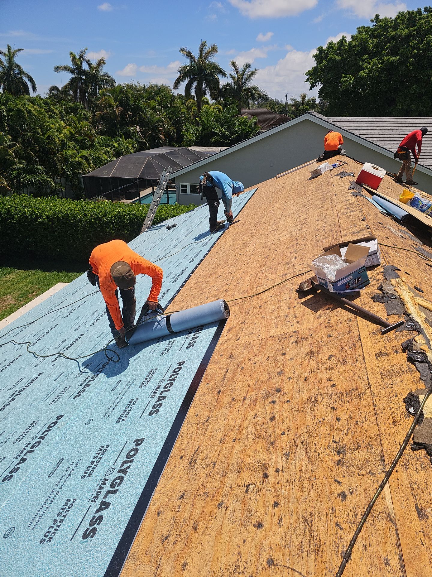 Workers installing blue roofing underlayment on a sloped roof beside exposed plywood, with trees and sky in back