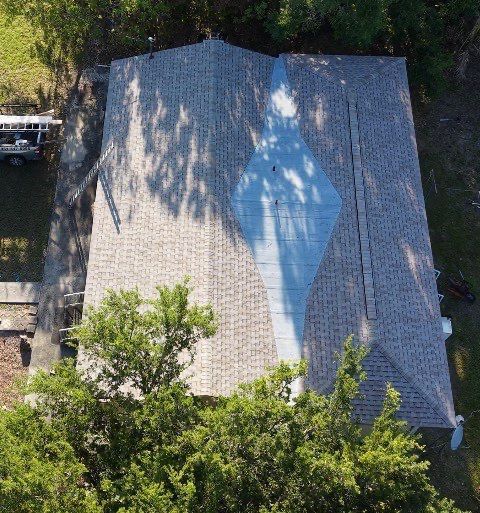 Aerial view of a house with a gray roof and blue tarp, surrounded by trees.