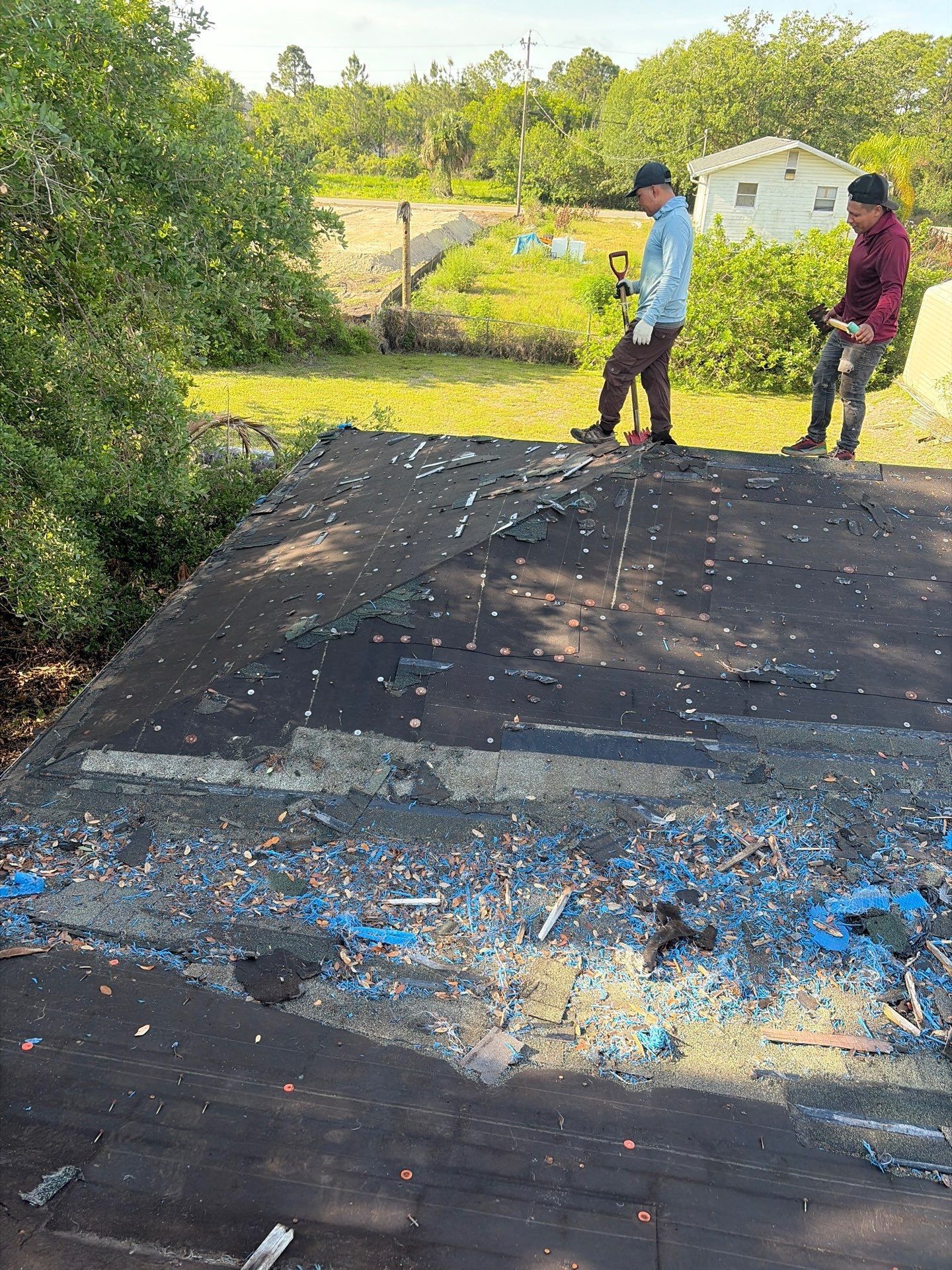 Two people standing beside a black tarp or roadway amid scattered blue debris in a sunny outdoor area