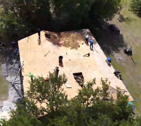 Aerial view of a beige rooftop with workers and surrounding trees, with a central opening and scattered materials.