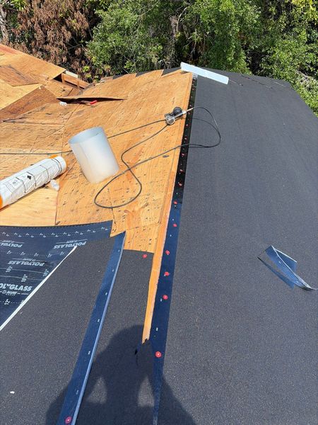 Roof under construction with exposed plywood, dark shingles, and tools near a tree line. Roof under construction with exposed plywood, dark shingles, and tools near a tree line.