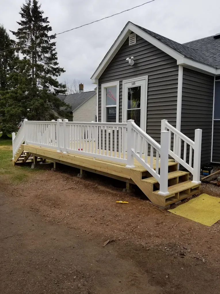White railing, wooden deck, and steps lead up to the entrance of a gray house with a cloudy sky background.