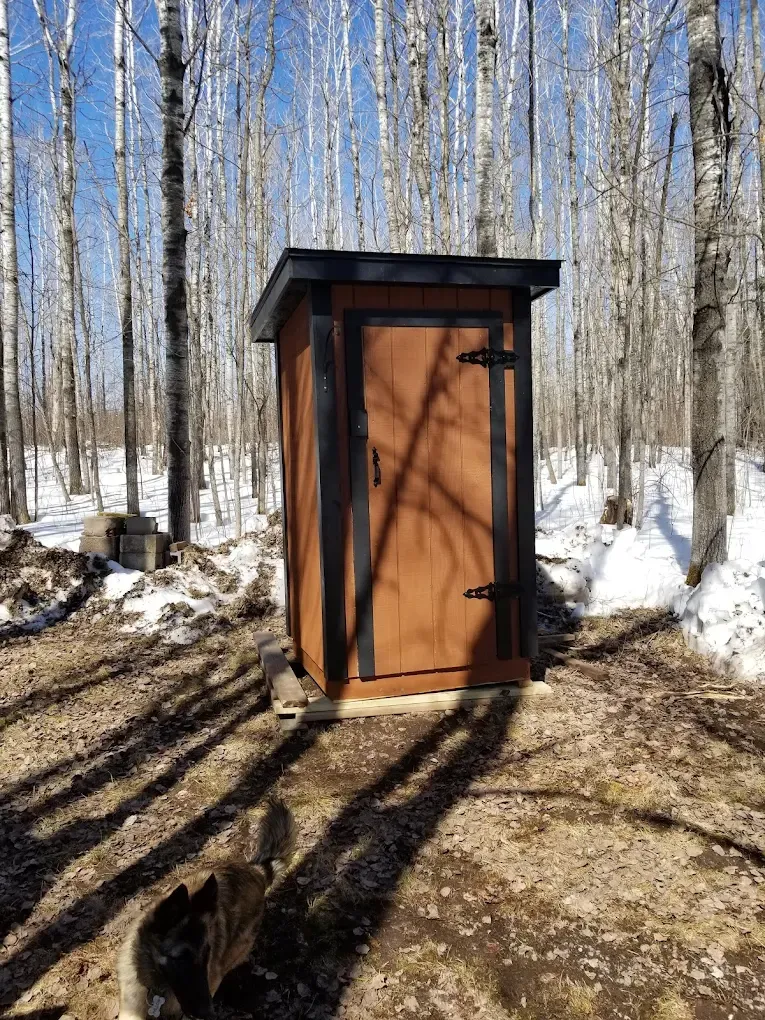 Brown outhouse with black trim stands in a snowy forest.