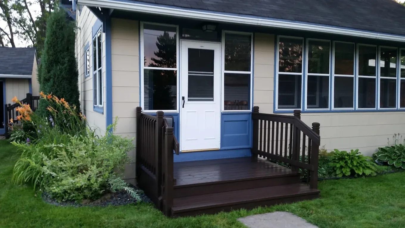 Brown porch with steps and railing in front of a small beige house. White door, blue paneling. Green grass.