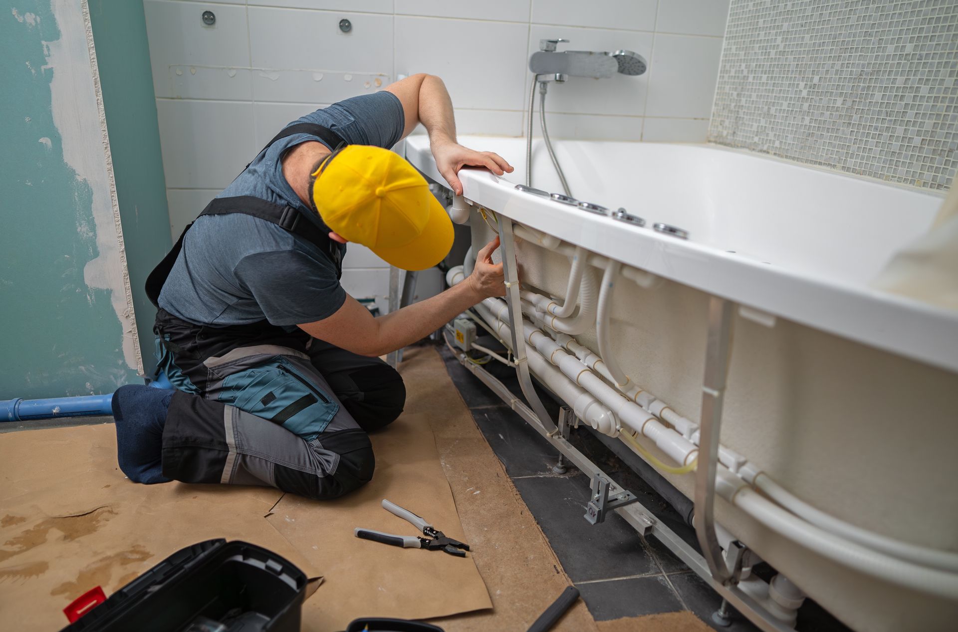Plumber kneels, working on bathtub plumbing in a bathroom under construction.