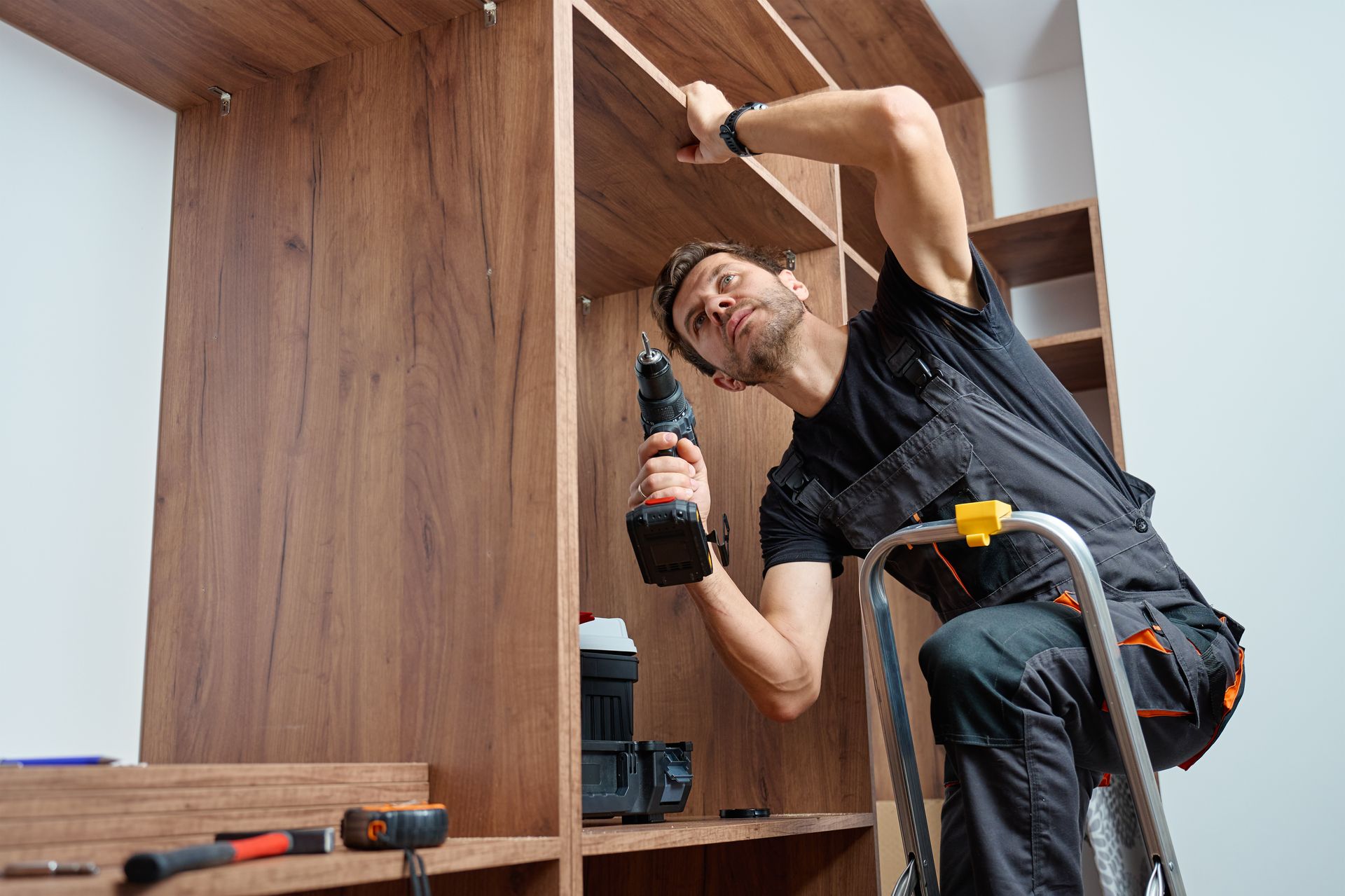 Man on a ladder, using a power drill to install shelving in a wooden cabinet.