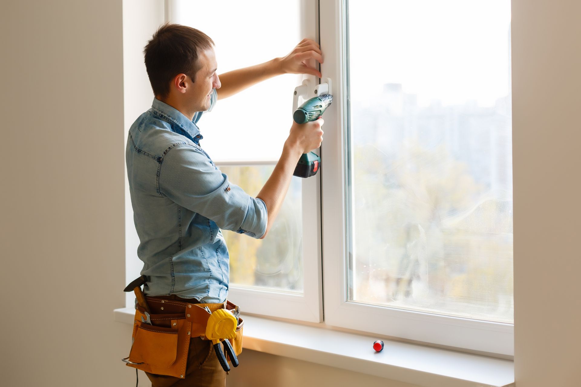 Man installing a window, using a drill, wearing a tool belt, in a white-walled room.
