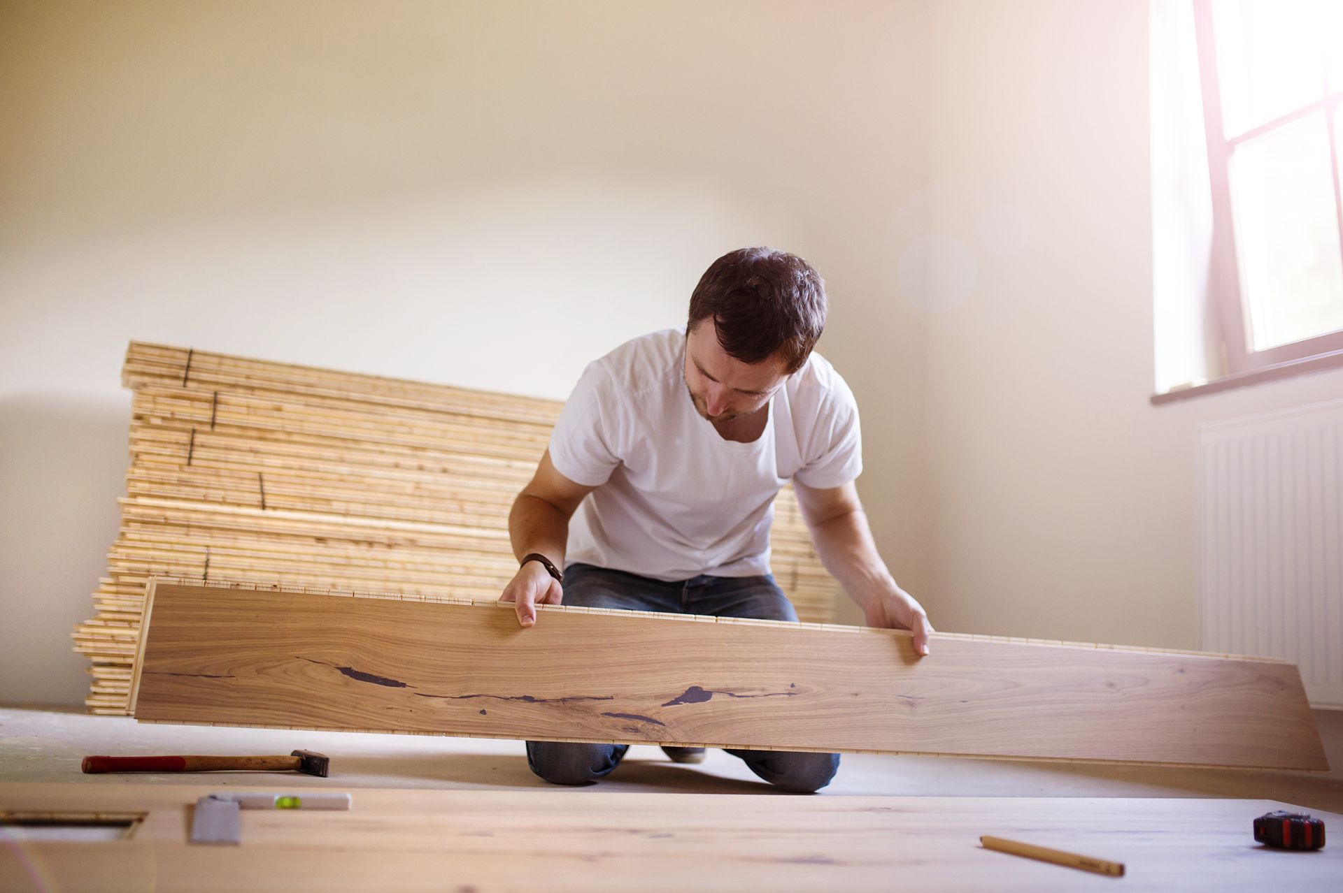 Man kneeling on floor, inspecting a wooden plank; a stack of planks behind him.