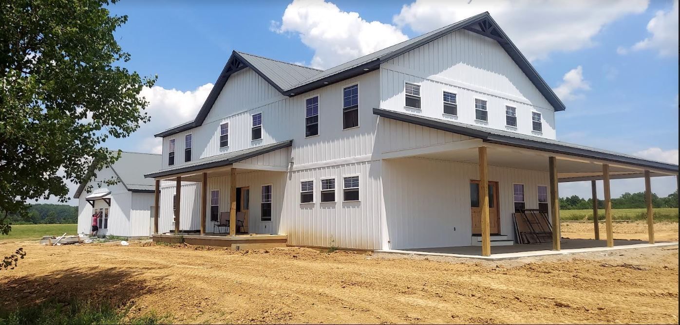 Large white house under construction with a covered porch and open dirt yard under a blue sky
