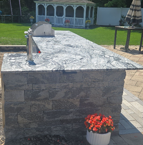 Outdoor stone grill or counter in a backyard patio, with red flowers in front and a gazebo in the background
