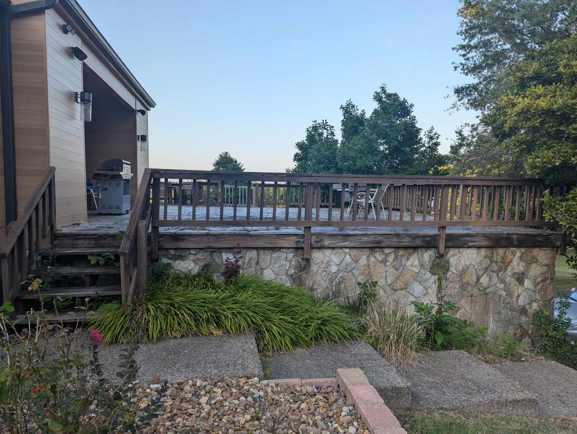 Stone patio with wooden deck railing beside a house, overlooking trees and gravel steps