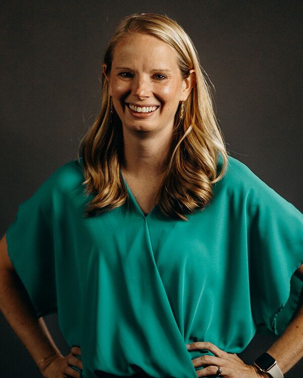 Woman in teal shirt smiles, hands on hips, against a dark gray background.