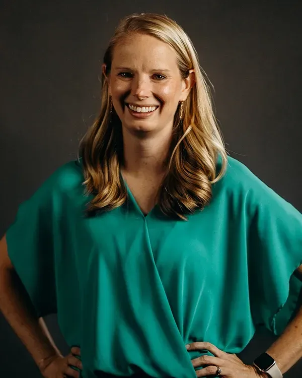 Woman with blonde hair wearing a teal blouse smiles, hands on hips, against a gray background.