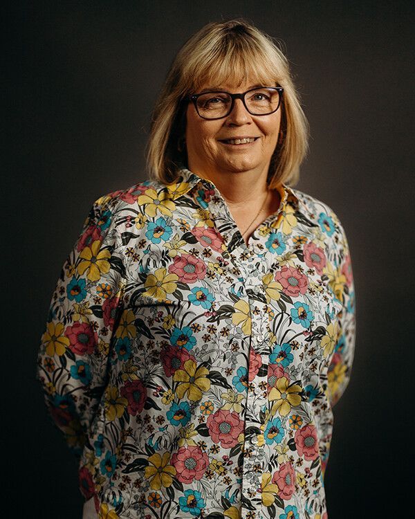 Woman with glasses smiling, wearing a floral-patterned shirt, posing against a dark background.
