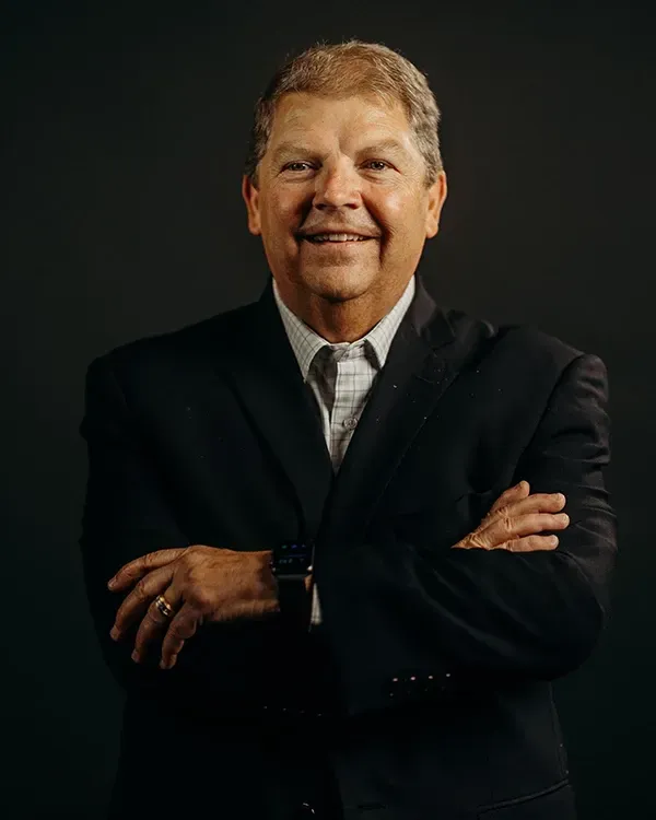 Man in suit, arms crossed, smiling, set against a dark background.